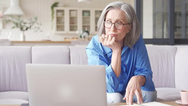 Middle aged senior older woman elearning online concept, watching business training class, live webinar on laptop computer remote working, social distance learning, writing notes from home office.
