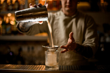 bartender holds sieve over old fashioned glass and pours cocktail from shaker.