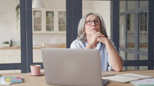 Thoughtful serious doubtful smart senior middle aged old woman looking away thinking of future vision, problem or question, business challenges, feeling doubt, sitting at home office work desk.