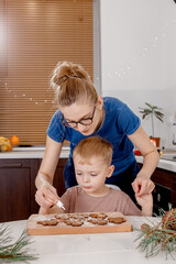 Young mother and son in kitchen making cookies. Christmas gingerbread man icing piping decoration. Woman with her child decorating traditional xmas cookie. Horizontal shot.