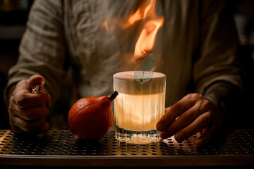 barman's hand holds old fashioned glass of cocktail with foam and with burning leaf.