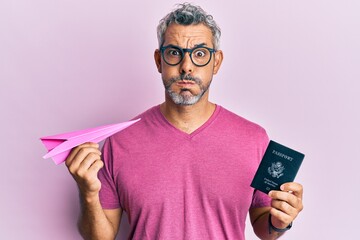 Middle age grey-haired man holding paper plane and passport puffing cheeks with funny face. mouth...