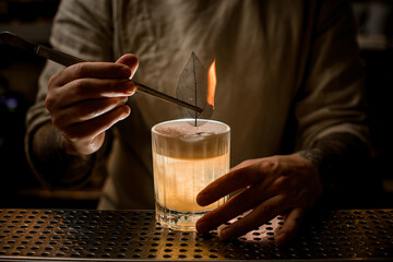 Fototapeta na wymiar man bartender holds tweezers with burning leaf to decorate glass of cocktail