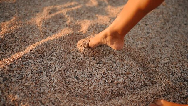 female leg foot of a young woman draws a heart on the sand and pebbles on the beach at sunset