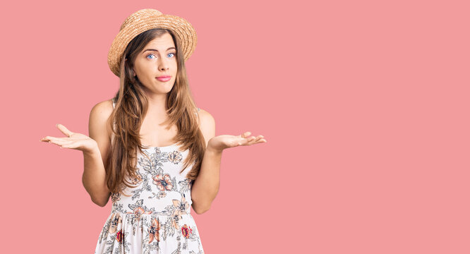 Beautiful caucasian young woman wearing summer hat clueless and confused expression with arms and hands raised. doubt concept.