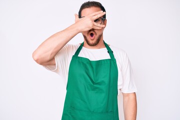 Fototapeta premium Young handsome man wearing apron peeking in shock covering face and eyes with hand, looking through fingers afraid