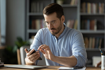 Serious businessman wearing spectacles using mobile phone sit at desk in cozy office. Manager solve issues with client remotely by modern wireless device, surfing web, learn new business app concept - obrazy, fototapety, plakaty