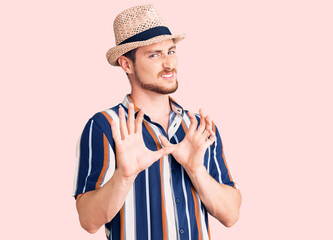 Young handsome caucasian man wearing summer hat disgusted expression, displeased and fearful doing disgust face because aversion reaction. with hands raised