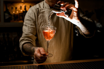 male bartender holds with hand wine glass with drink and adds liquid from jigger