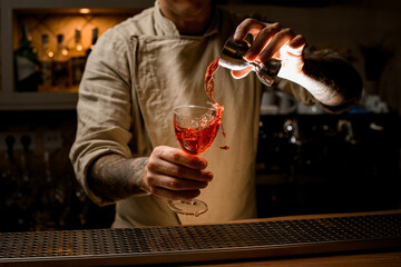 Man at bar holds cocktail glass with splashing alcoholic drink and adds liquid from jigger