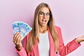 Young blonde woman wearing business style holding south african rands banknotes celebrating achievement with happy smile and winner expression with raised hand