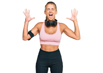 Young blonde woman wearing gym clothes and using headphones celebrating mad and crazy for success with arms raised and closed eyes screaming excited. winner concept