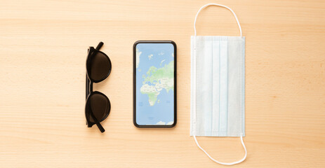 Travel concept, a face mask, sunglasses and a smartphone with a World Map are arranged on a wooden table. In the context of the COVID-19 pandemic, CDC recommends that everyone wear a mask.
