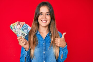 Beautiful caucasian woman holding australian dollars smiling happy and positive, thumb up doing excellent and approval sign