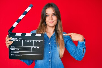 Beautiful caucasian woman holding video film clapboard pointing finger to one self smiling happy...
