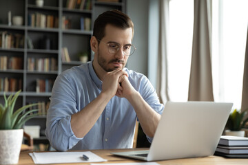 Young businessman sit at home office desk looks at laptop read media news online, learn new e app, analyzing project, stuck with challenge business task, thinks over problem, search solution concept