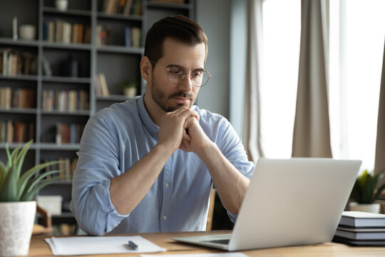 Young Businessman Sit At Home Office Desk Looks At Laptop Read Media News Online, Learn New E App, Analyzing Project, Stuck With Challenge Business Task, Thinks Over Problem, Search Solution Concept