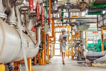 Full length of hardworking blond female supervisor in suit with helmet and face mask on holding tablet and walking around heating plant during corona virus outbreak.