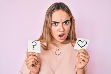 Young beautiful blonde woman holding heart and question mark reminder in shock face, looking...