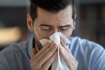 Close up young 35s man in glasses holding tissue and sneezing, blowing runny nose in paper...