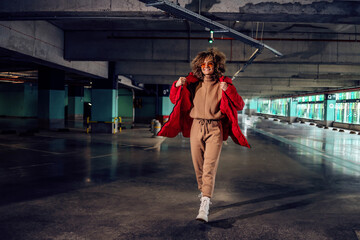 Young fashionable woman with curly hair holding hand in pocket of jacket and walking in underground...