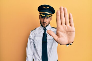 Handsome hispanic man wearing airplane pilot uniform doing stop sing with palm of the hand. warning...