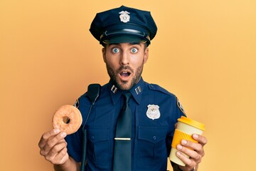 Handsome hispanic police man eating donut and drinking coffee afraid and shocked with surprise and...
