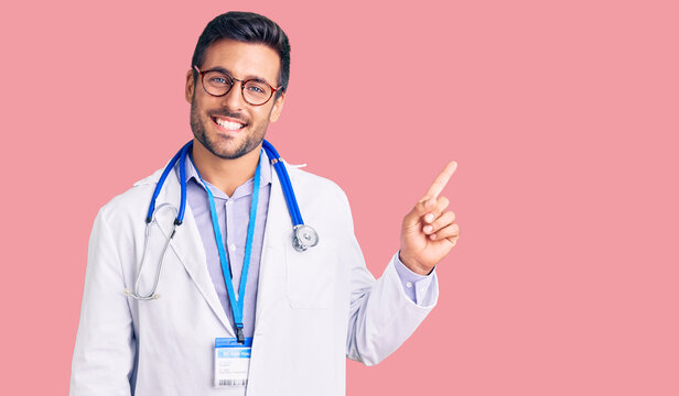 Young hispanic man wearing doctor uniform and stethoscope with a big smile on face, pointing with hand finger to the side looking at the camera.