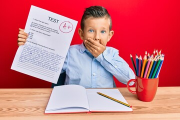 Adorable caucasian kid holding passed test sitting on the table covering mouth with hand, shocked...