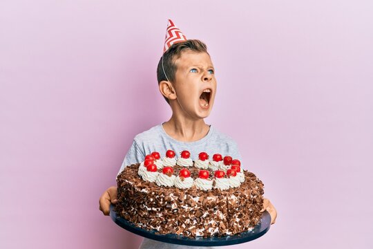 Adorable caucasian kid celebrating birthday with cake angry and mad screaming frustrated and furious, shouting with anger. rage and aggressive concept.