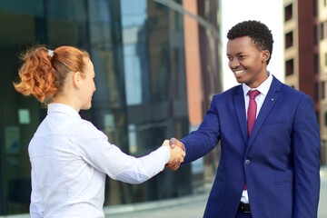 Two happy smiling people: businessman and businesswoman are shaking hands, greeting. White European young woman and black African Afro American man in formal suit outdoors, looking at camera