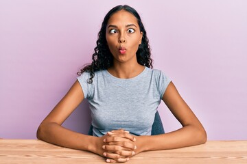 Young african american girl wearing casual clothes sitting on the table making fish face with lips, crazy and comical gesture. funny expression.