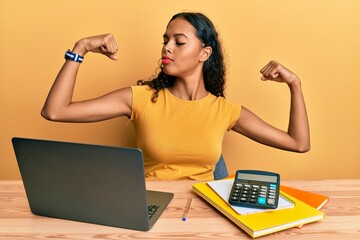 Young african american girl working at the office with laptop and calculator showing arms muscles...