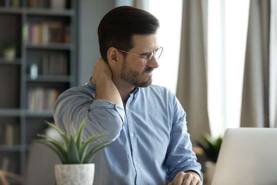 Young man sit at workplace desk touch neck feeling ache, massaging nape to relief painful feelings. Poor sitting posture during laptop usage, sedentary lifestyle, chronic disorder fibromyalgia concept
