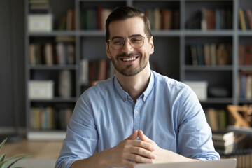 Happy millennial businessman smile look at camera sit at desk in home office. Concept of video call webinar participant, successful entrepreneur, company owner, teacher or business trainer portrait