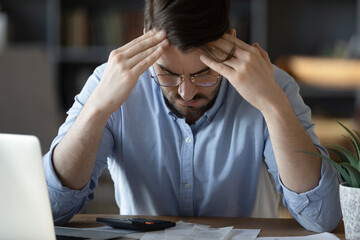 35s businessman sitting at workplace desk with bowed head, on table lot of unpaid receipts, overdue...