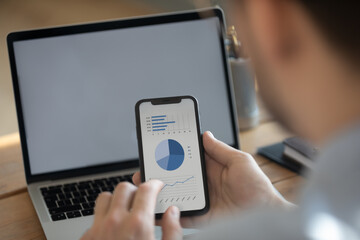 Businessman holding smart phone with financial data blue graphs charts on screen, laptop empty monitor with mock up, close up view over man shoulder. Synchronize information, new app, software concept