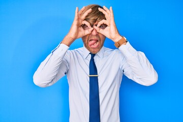 Young blond man wearing business clothes and glasses doing ok gesture like binoculars sticking tongue out, eyes looking through fingers. crazy expression.