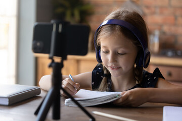 Smart lessons. Cute little girl in headphone set engaged in study online using mobile phone fixed on support, watching video course by internet, handwriting taking notes, thinking on interactive task