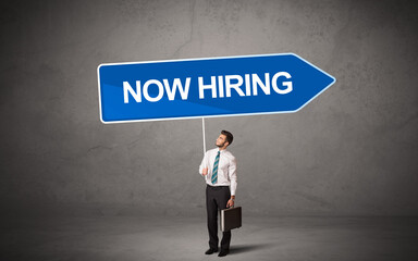 Young business person in casual holding road sign with NOW HIRING inscription, new business direction concept
