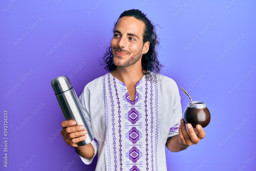 Sticker young handsome man with long hair drinking mate infusion smiling looking to the side and staring awa