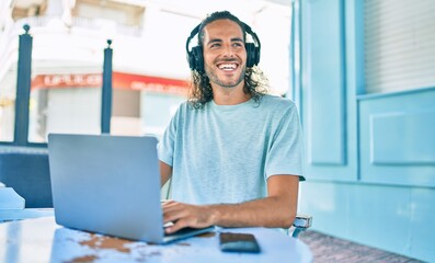 Young hispanic man smiling happy working using laptop and headphones at terrace of coffee shop.