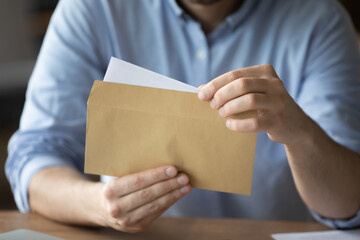 Businessman sitting at workplace desk holding yellow envelope take out received paper letter notice sheet, close up image. Business correspondence, corporate news, notification or invitation concept