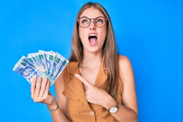 Young blonde girl holding south african rand banknotes angry and mad screaming frustrated and furious, shouting with anger looking up.