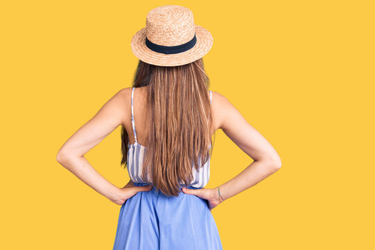 Young beautiful blonde woman wearing summer hat standing backwards looking away with arms on body