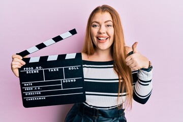 Young beautiful redhead woman holding video film clapboard smiling happy and positive, thumb up...