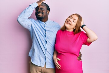 Young interracial couple expecting a baby, touching pregnant belly smiling confident touching hair with hand up gesture, posing attractive and fashionable