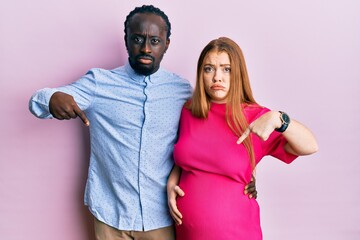 Obraz premium Young interracial couple expecting a baby, touching pregnant belly pointing down looking sad and upset, indicating direction with fingers, unhappy and depressed.