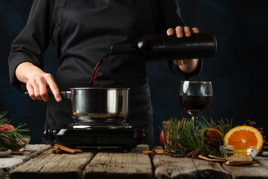 Close-up View Of Chef Pours Red Wine Into The Pot For Preparing Mulled Wine On Rustic Wooden Table With Festive Composition Background. Backstage Of Cooking Hot Drink With Spices. Frozen Motion.