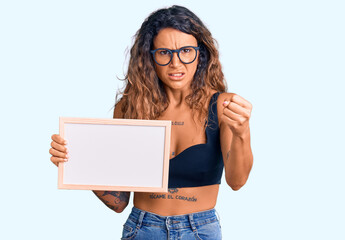Young hispanic woman with tattoo holding empty white chalkboard annoyed and frustrated shouting with anger, yelling crazy with anger and hand raised